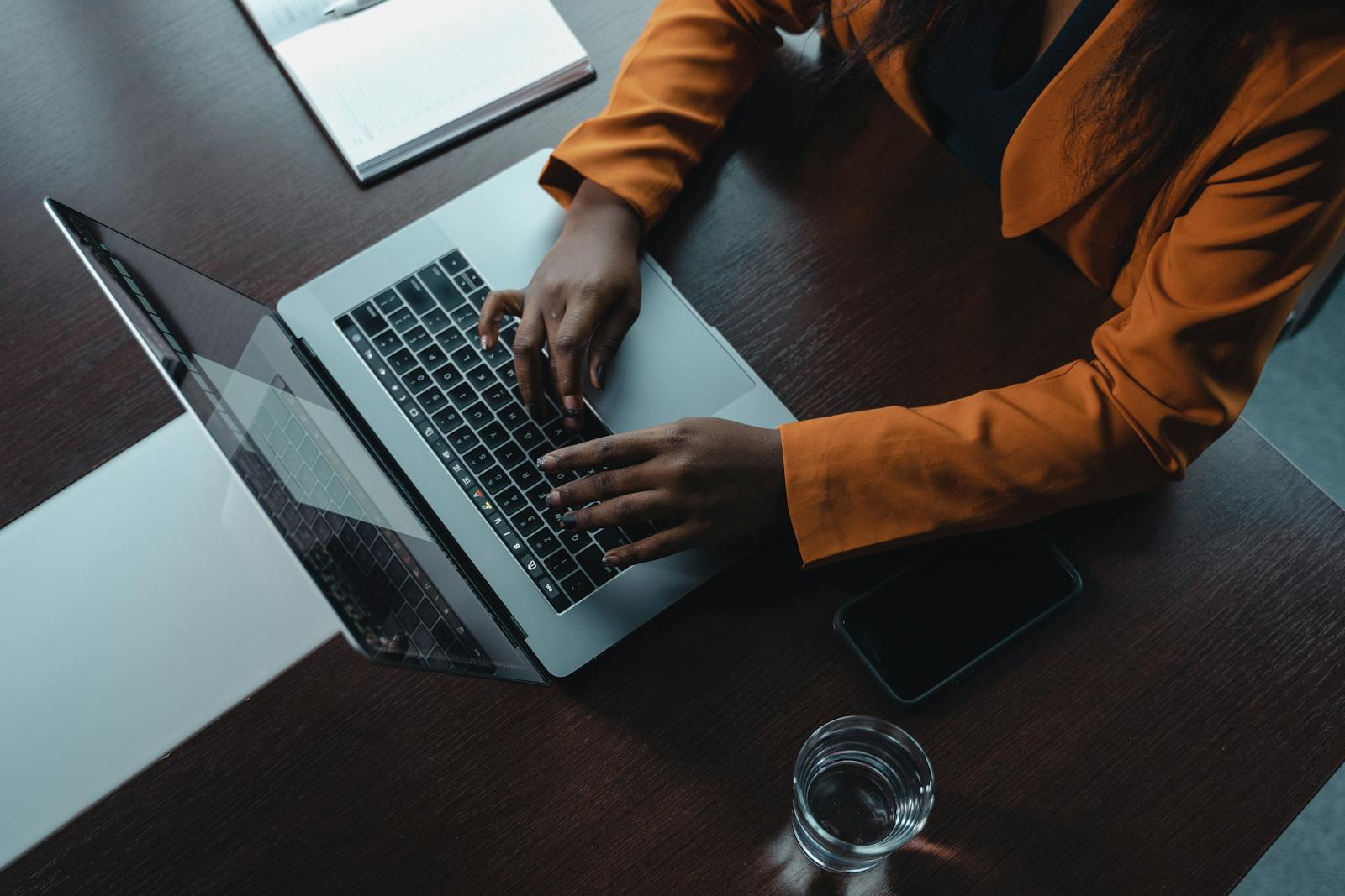 A woman in an orange blazer typing on a laptop in an office setting.